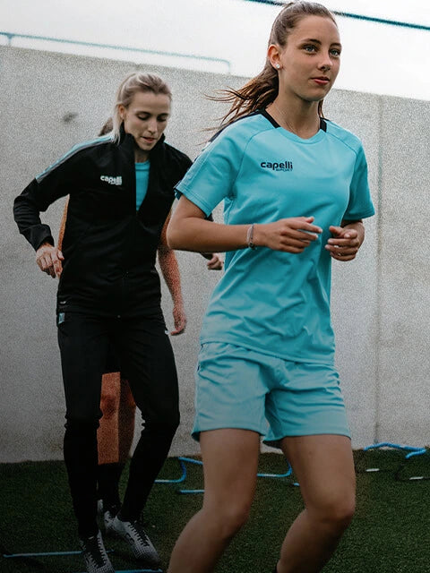 Two women running on a sports field, wearing Capelli sports jerseys.