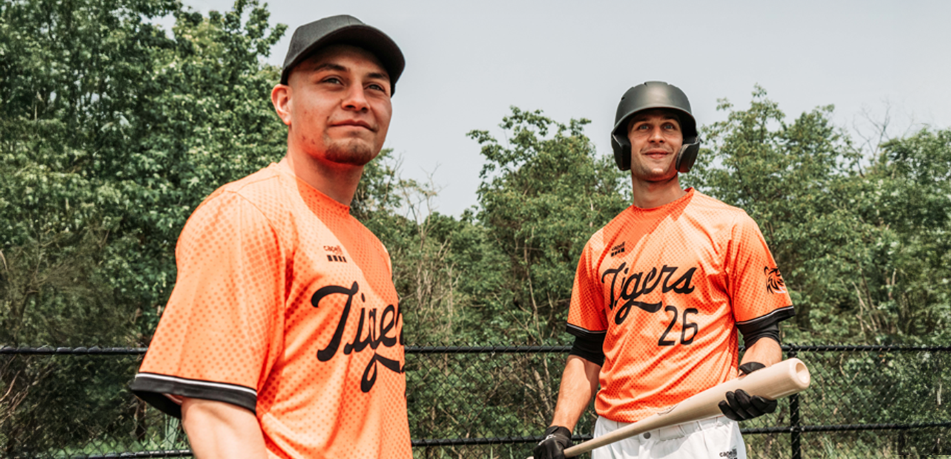 Two baseball players in orange 'Tigers' jerseys standing outdoors with trees in the background.