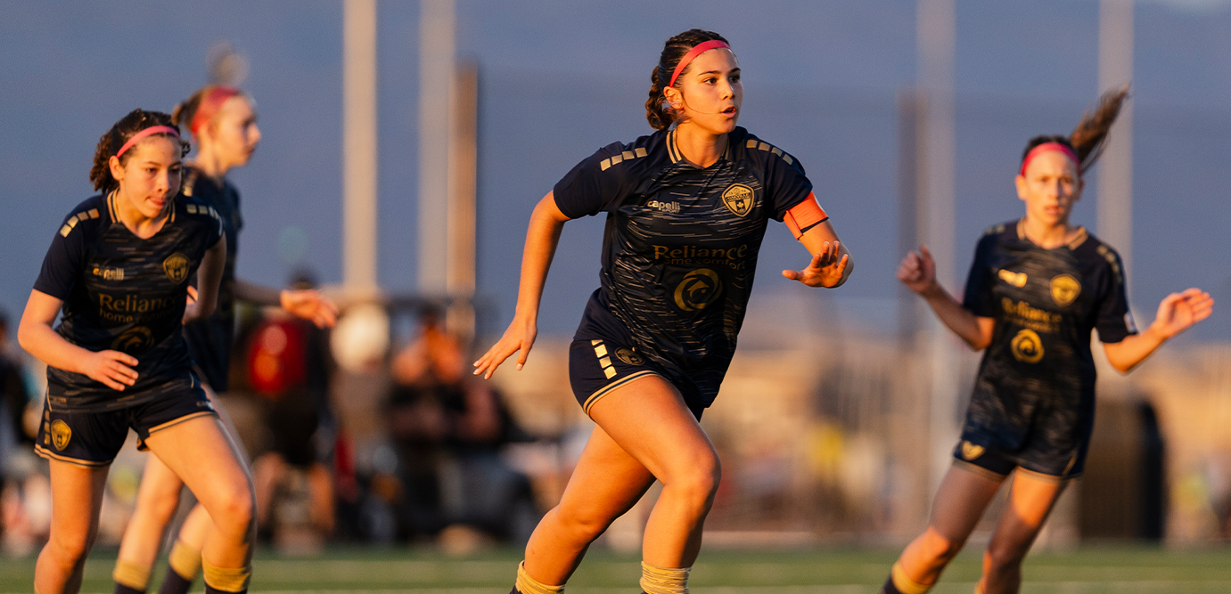 Three female soccer players in matching dark navy jerseys and shorts running on a field during a game.