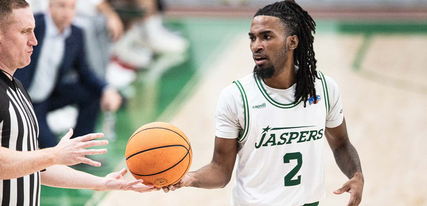 A basketball player in a white and green Jaspers jersey with the number 2 holds an orange basketball while interacting with a referee on a court.