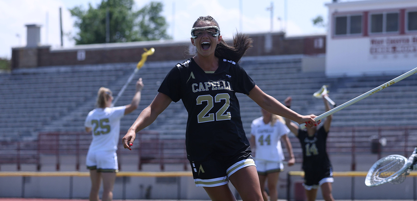 A female lacrosse player wearing a black and gold Capella jersey with the number 22, celebrating on an outdoor sports field with stadium bleachers in the background.