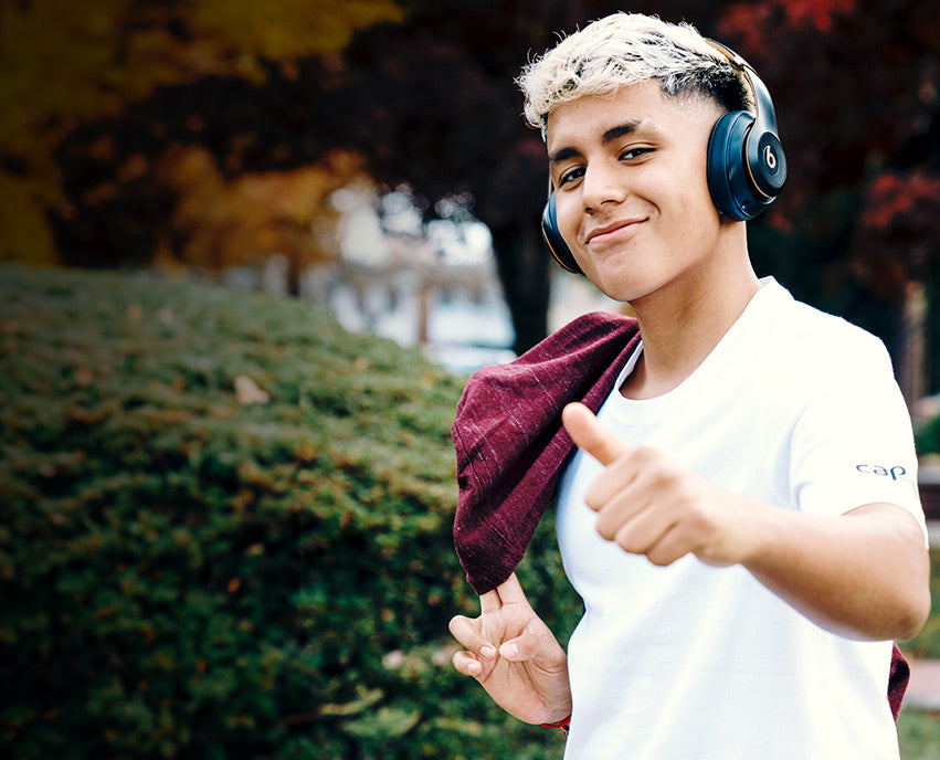 Person wearing headphones and holding a maroon hoodie, giving a thumbs-up outdoors.